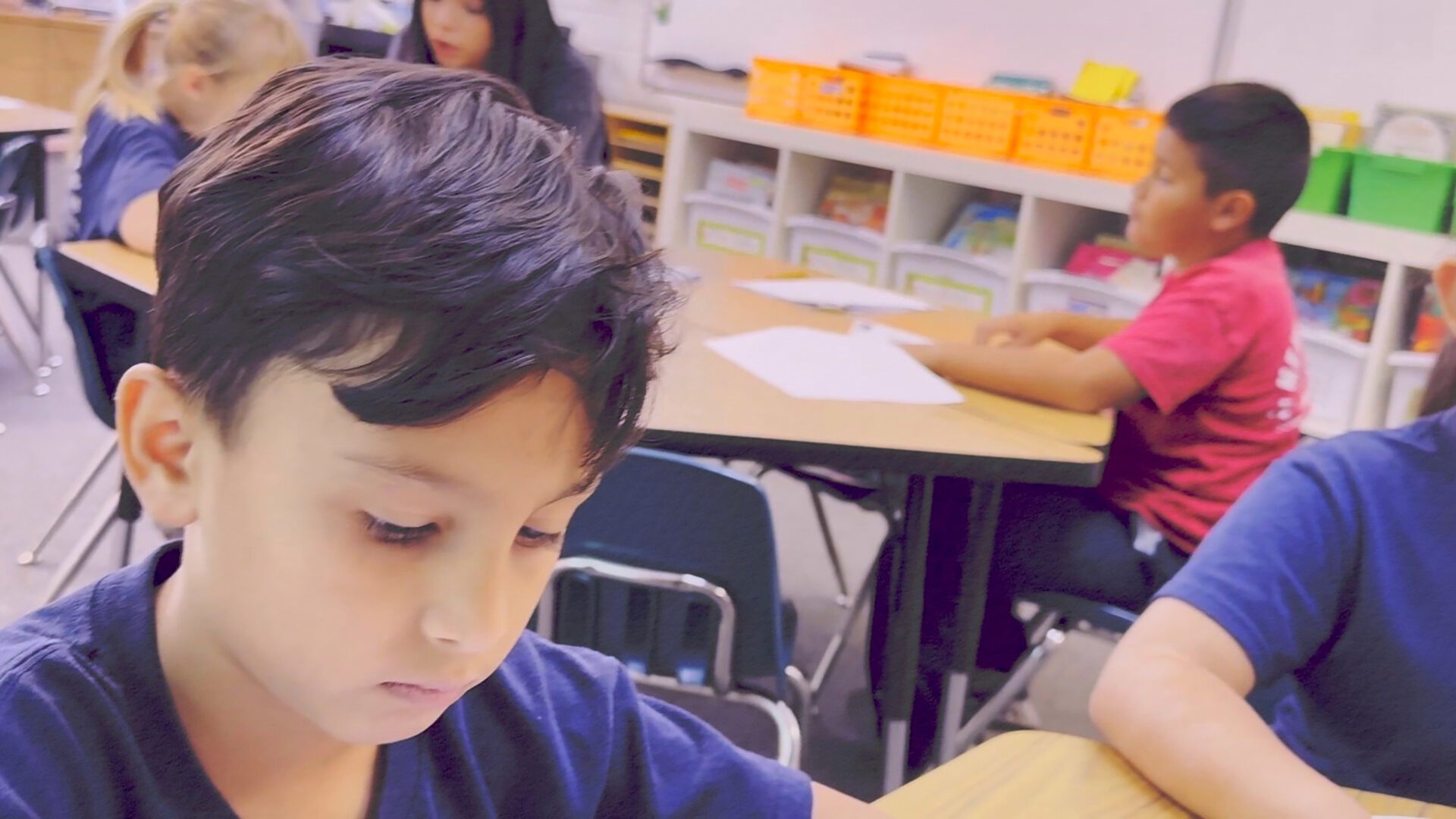 -sm-students-title-pic-no-text-larger-2a A young boy focused on writing or drawing at a classroom desk with colorful storage bins in the background.